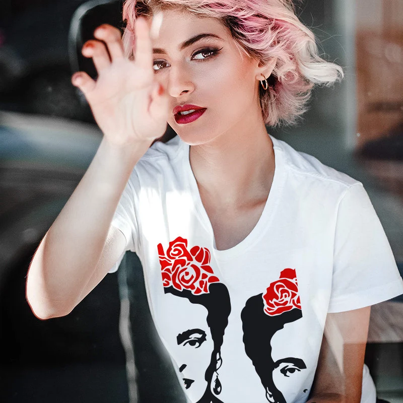 Fit short hair blond woman with sligh pinkish hair wearing a White t-shirt. The design of the -t-shirt is a contour face of frida khalo in black, with red roses on top of hair. The face is split in half verticaly, the two parts of design are swiched with space between them. the left part is slightily moved up. Design by Correct Studio